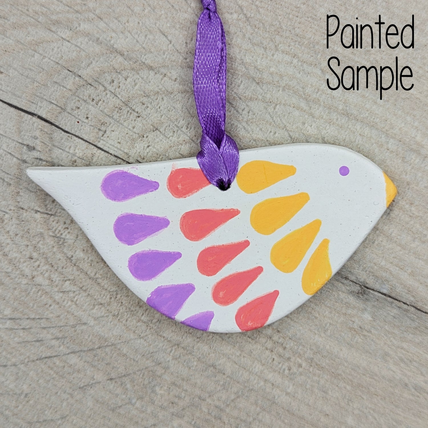 painted stylised bird clay decoration, painted in yellow, pink and lilac, with a purple hanging ribbon