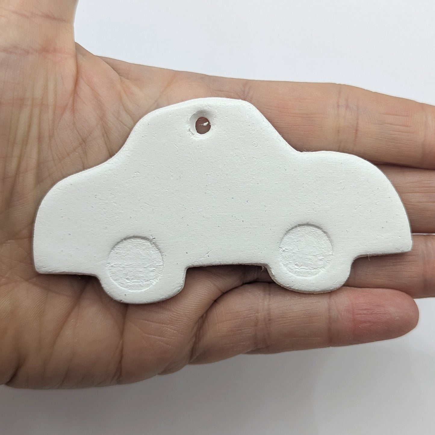 White clay car shaped object, with car wheels stamped into the clay, held in the palm of hand to show scale