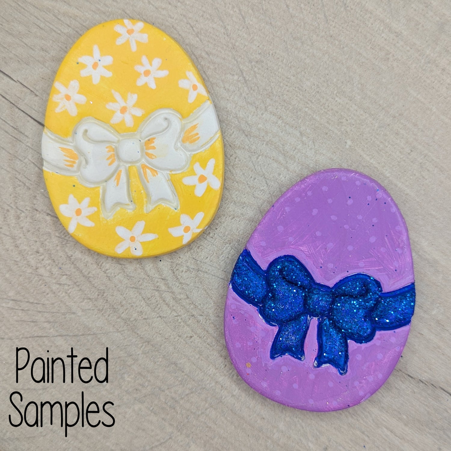 Two painted easter egg shaped decorations, one yellow with white flowers and a bow, the other purple with a blue bow