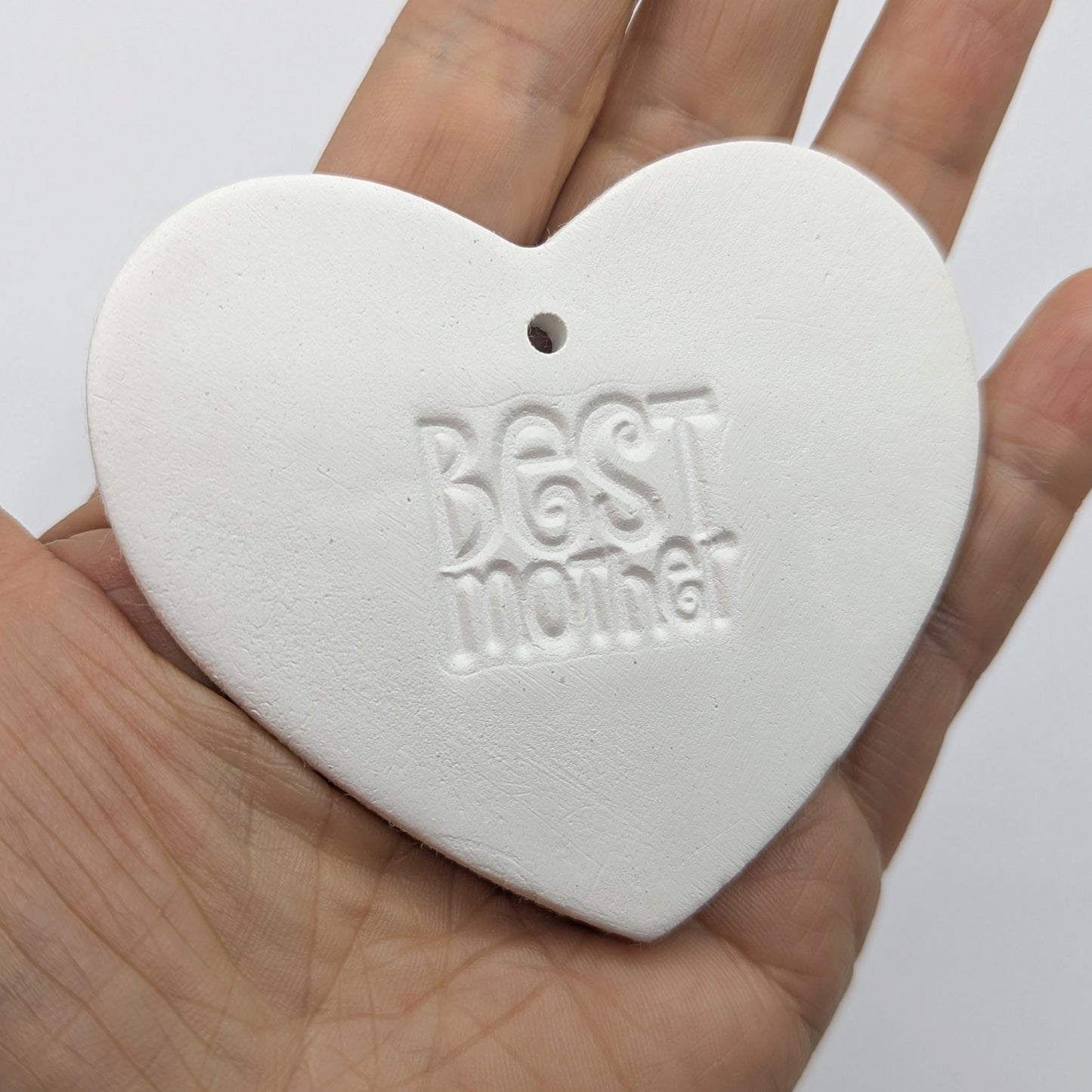 White heart shaped air dried clay tag with 'Best Mother' stamped into the clay, held in a hand against a white background