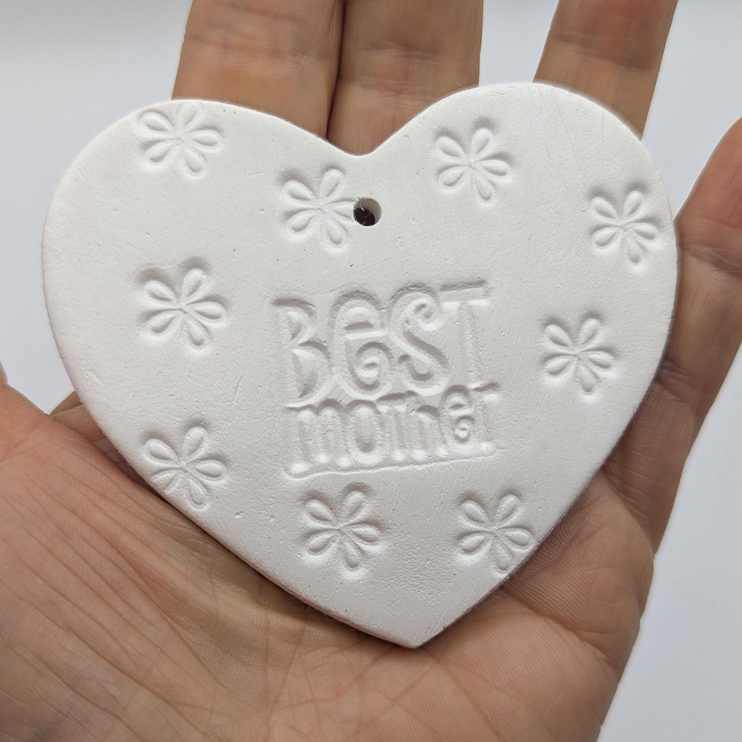 White heart shaped air dried clay tag with 'Best Mother' and small cute flowers stamped into the clay, held in a hand against a white background