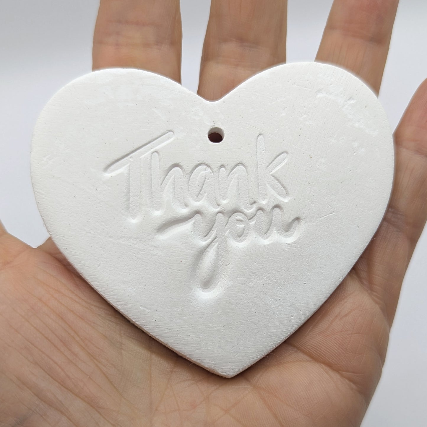 White heart shaped air dried clay tag with 'Thank you' stamped into the clay, held in a hand against a white background