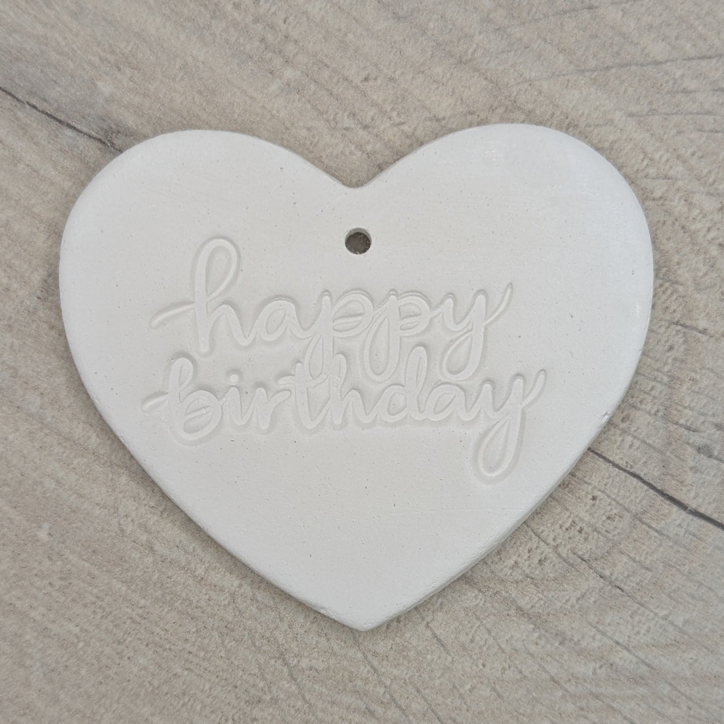 Heart shaped air dried clay decoration / tag with 'happy birthday' in lower case stamped into the clay, on a wooden background