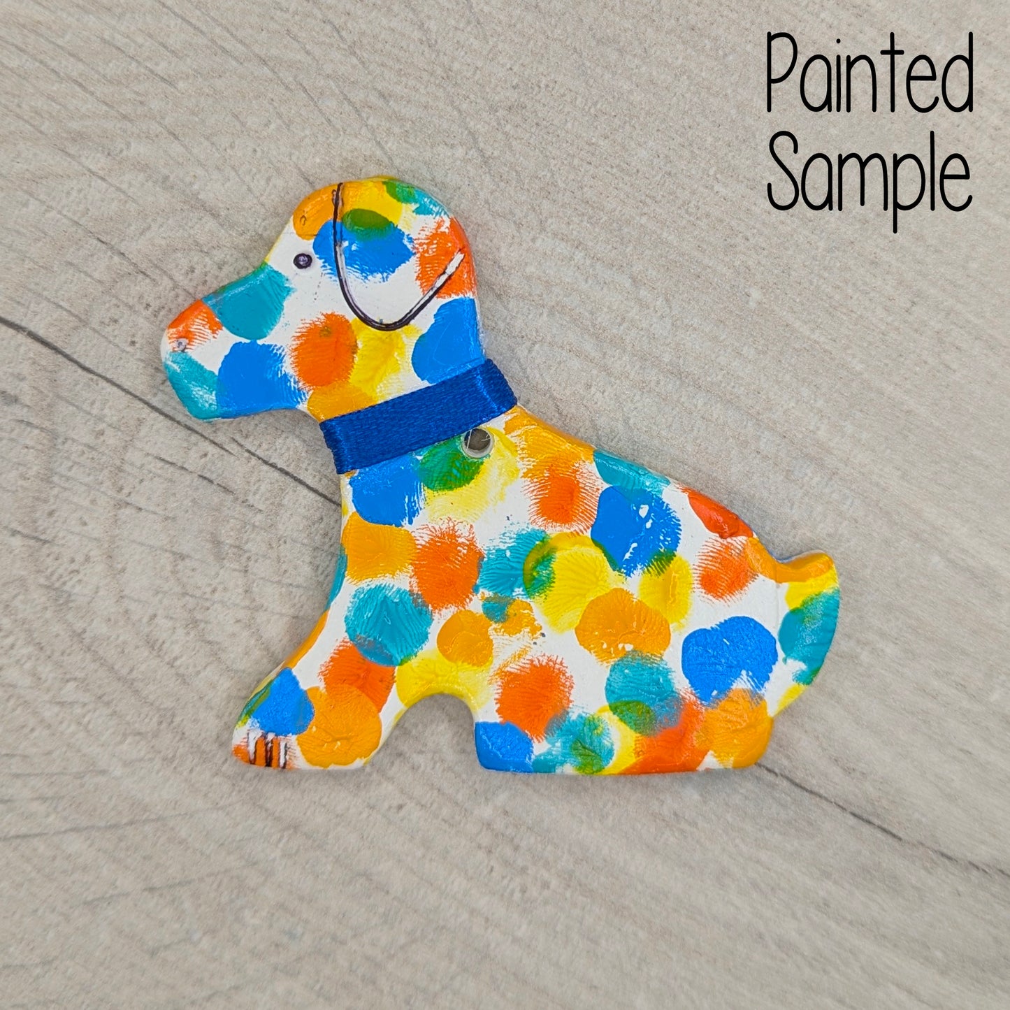 Colorful dog-shaped clay object with a blue collar on a wooden surface, labeled 'Painted Sample'.