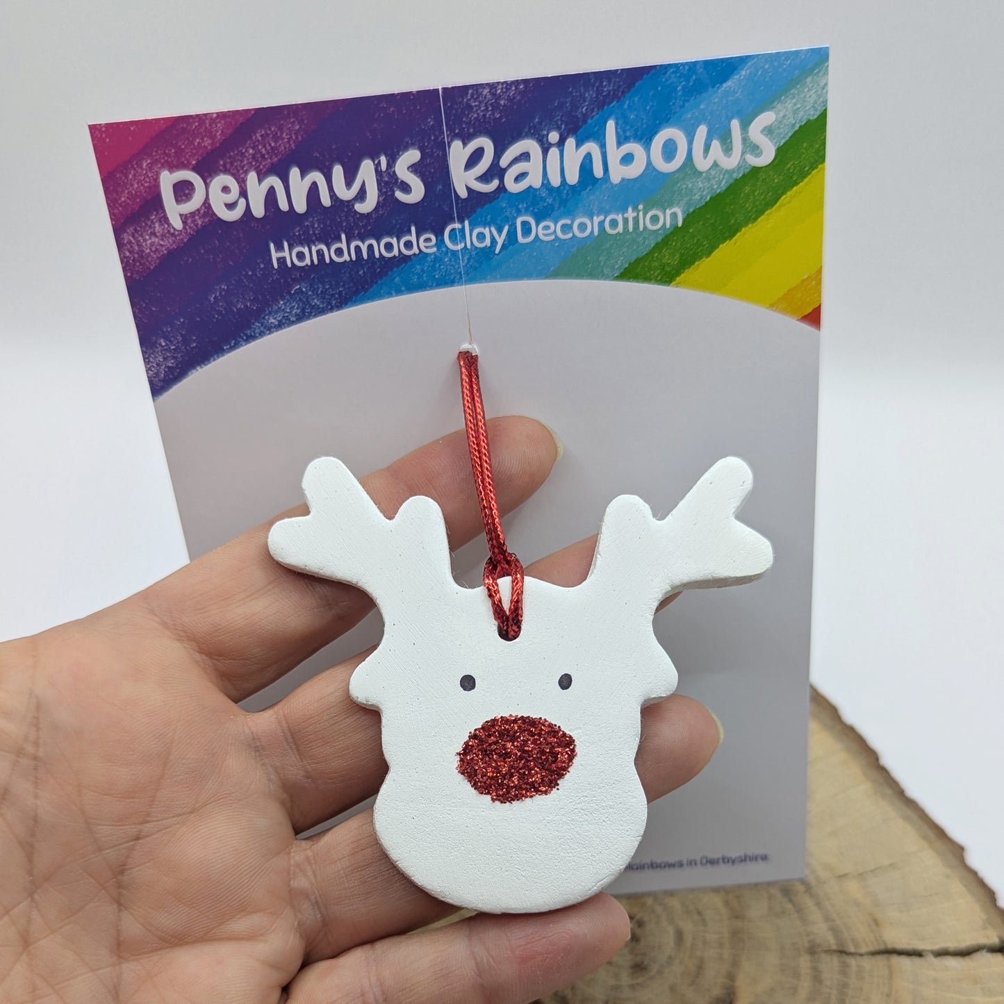 White clay reindeer ornament with red glitter nose and cord held in front of 'Penny's Rainbows' packaging