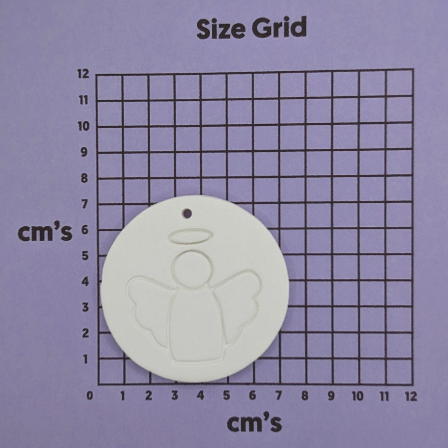 White clay disc with a simple clean angel design stamped onto it, shown on a purple cm size grid for scale