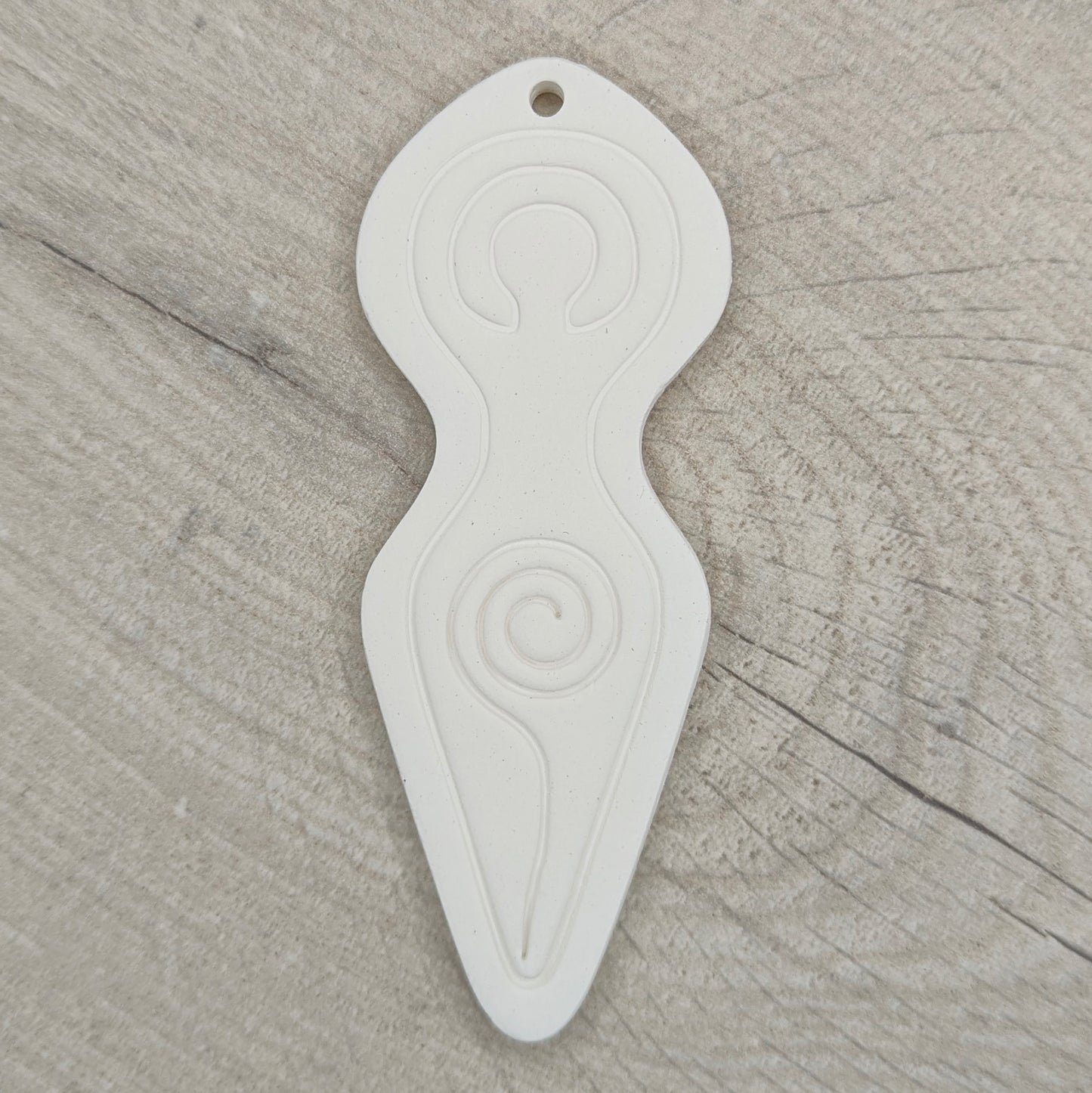 Spiral goddess shaped clay decoration with a hanging hole above her head area, shown on a light wood background