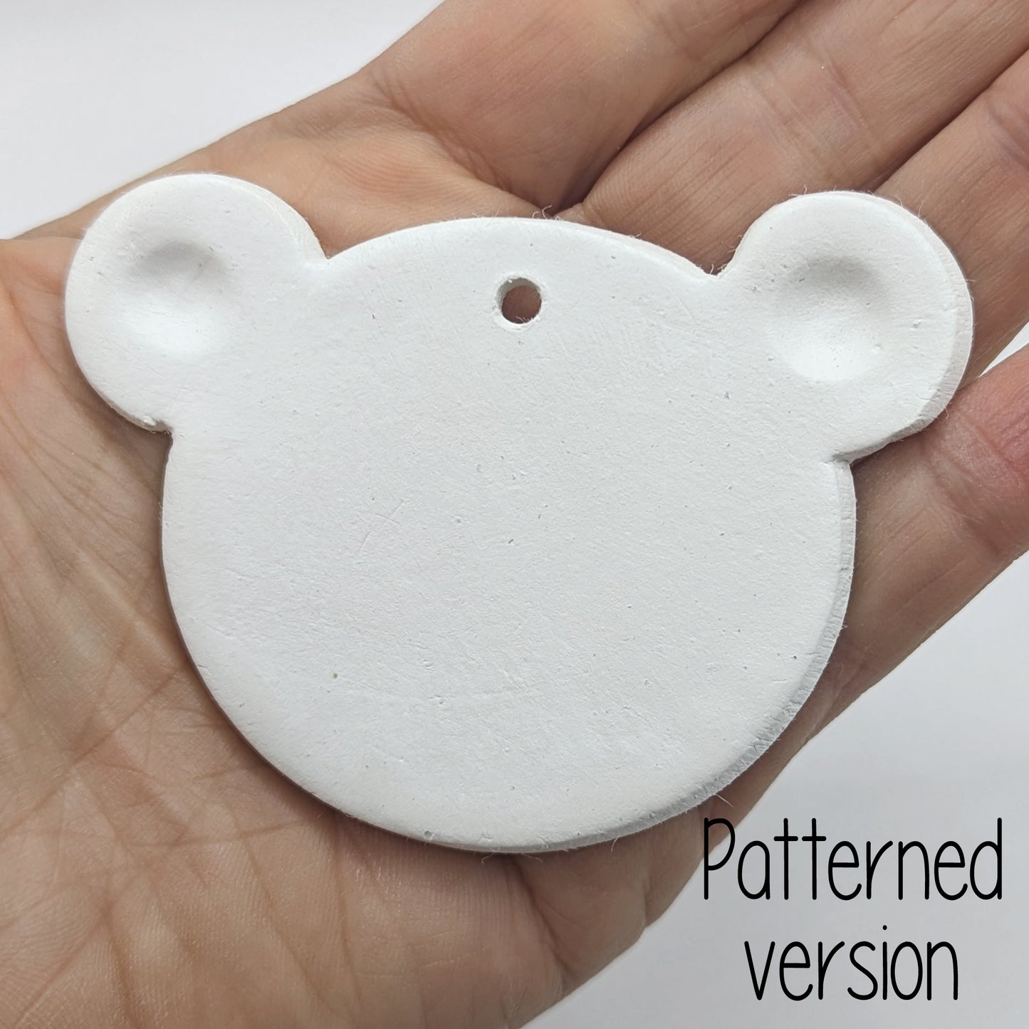 White clay decoration in the shape of a teddy bear head with ears stamped in, held in hand to show scale