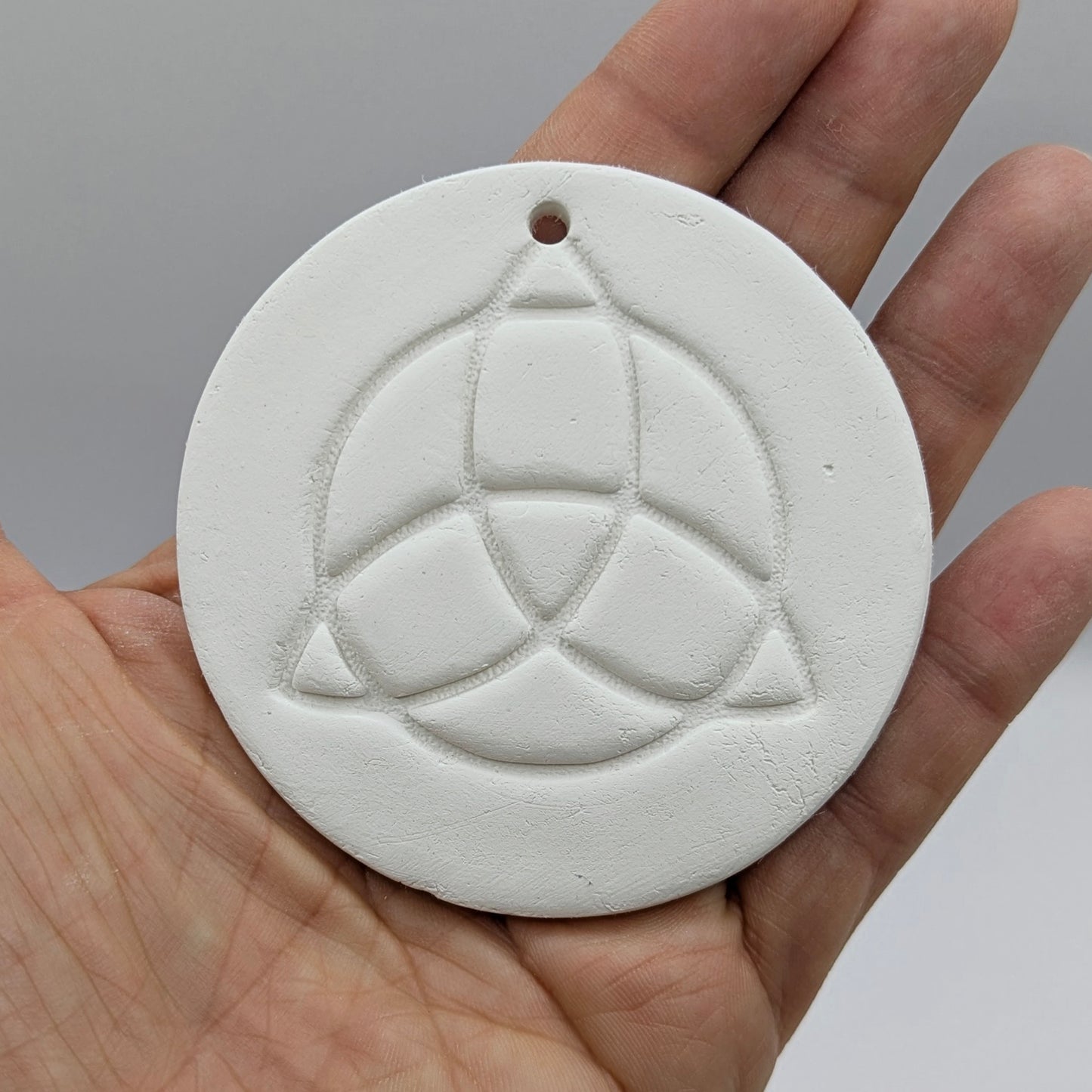 white air dried clay decoration with the triquetra symbol stamped into it held in the palm of some ones hand to show scale