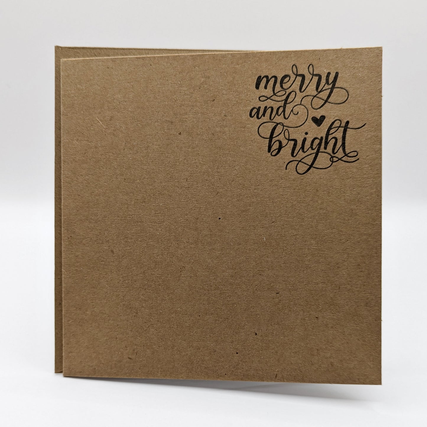 close up of wording 'merry and bright' in a script font, stamped in black ink onto a kraft card comes with matching envelope
