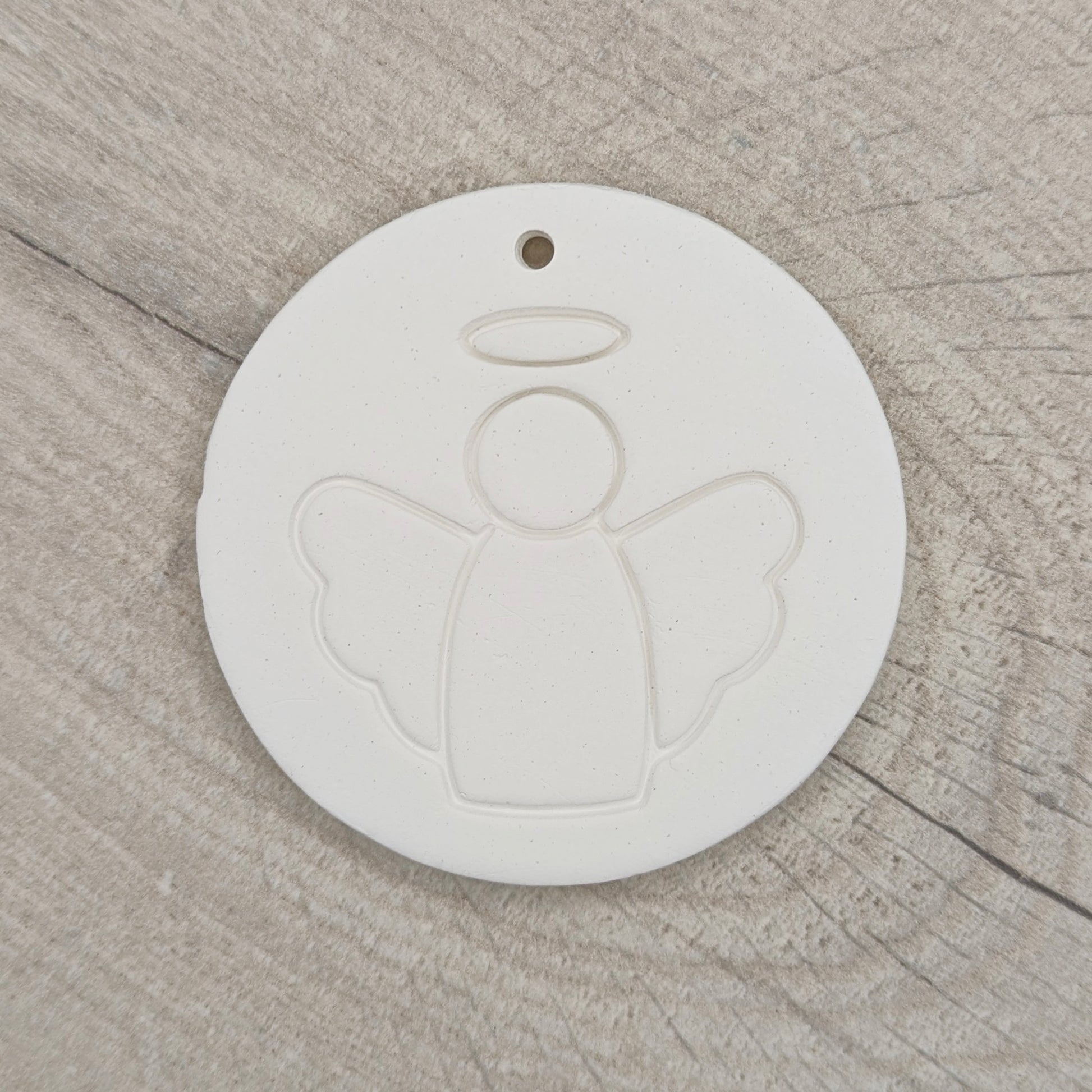 White clay disc with a simple clean angel design stamped onto it, hanging hole in the top, shown on a wooden background