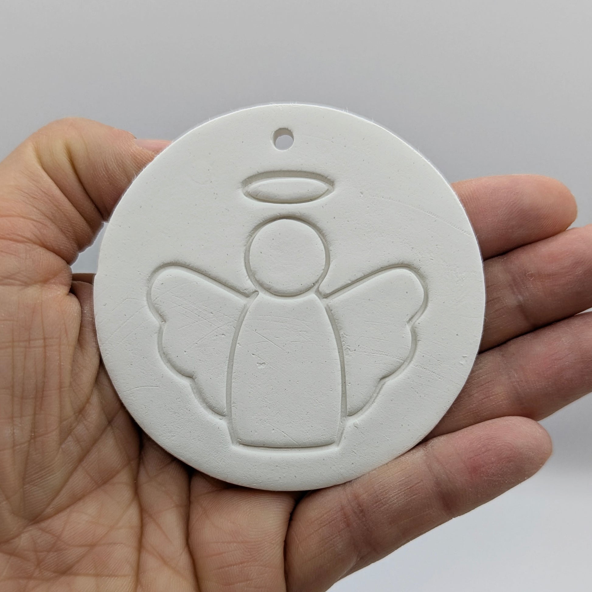 White clay disc with a simple clean angel design stamped onto it, shown in some ones hand for scale