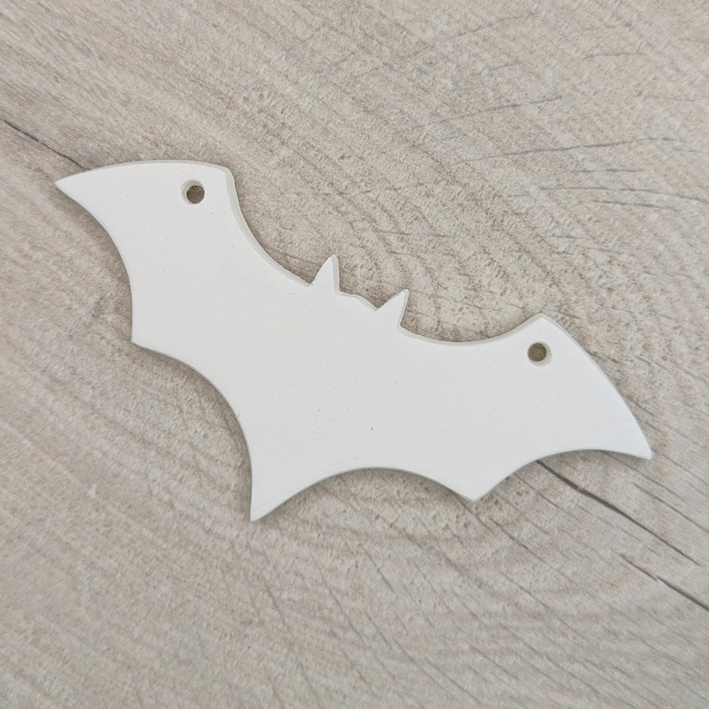 white air dried clay decoration in the shape of a bat with a hanging hole in each wind to hang it up, shown on a light wooden background