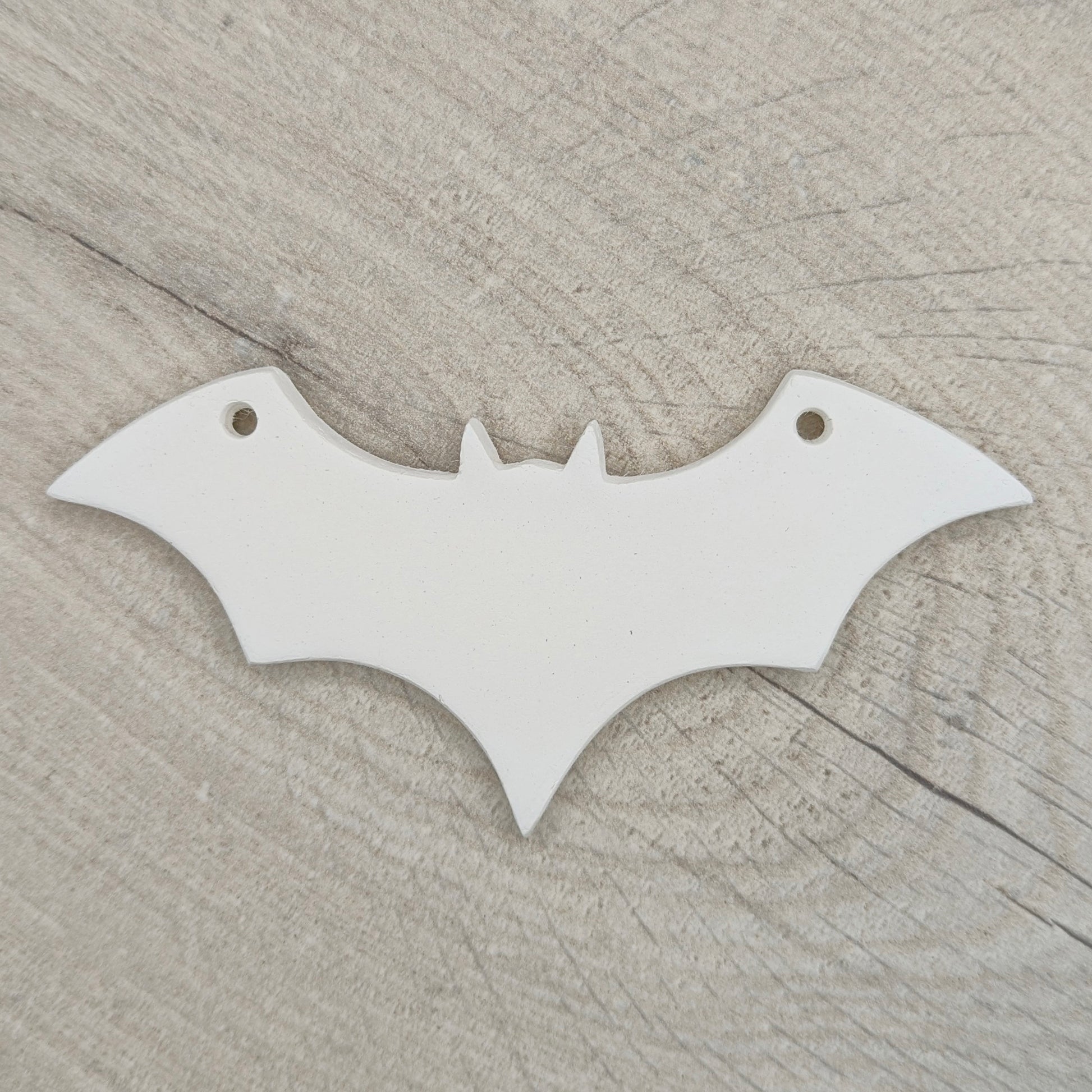 white air dried clay decoration in the shape of a bat with a hanging hole in each wind to hang it up, shown on a light wooden background