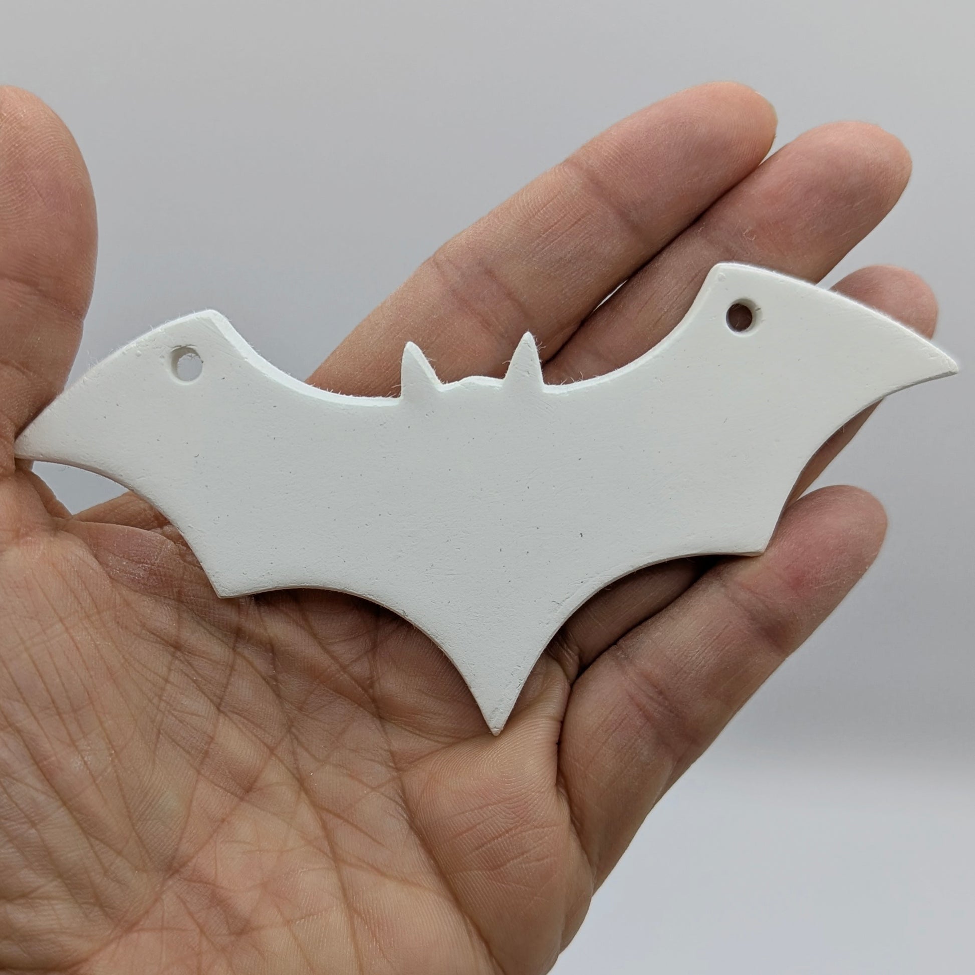 white air dried clay decoration in the shape of a bat with a hanging hole in each wind to hang it up, shown in some ones hand for scale
