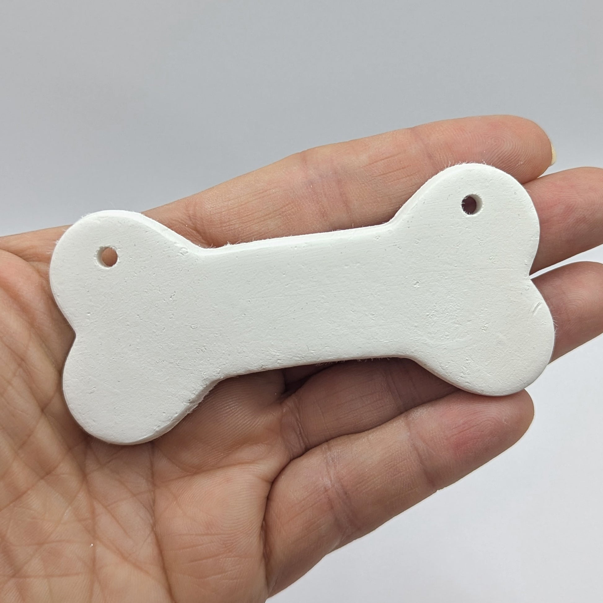 Bone shaped white clay decoration  held in hand for scale, two hanging holes