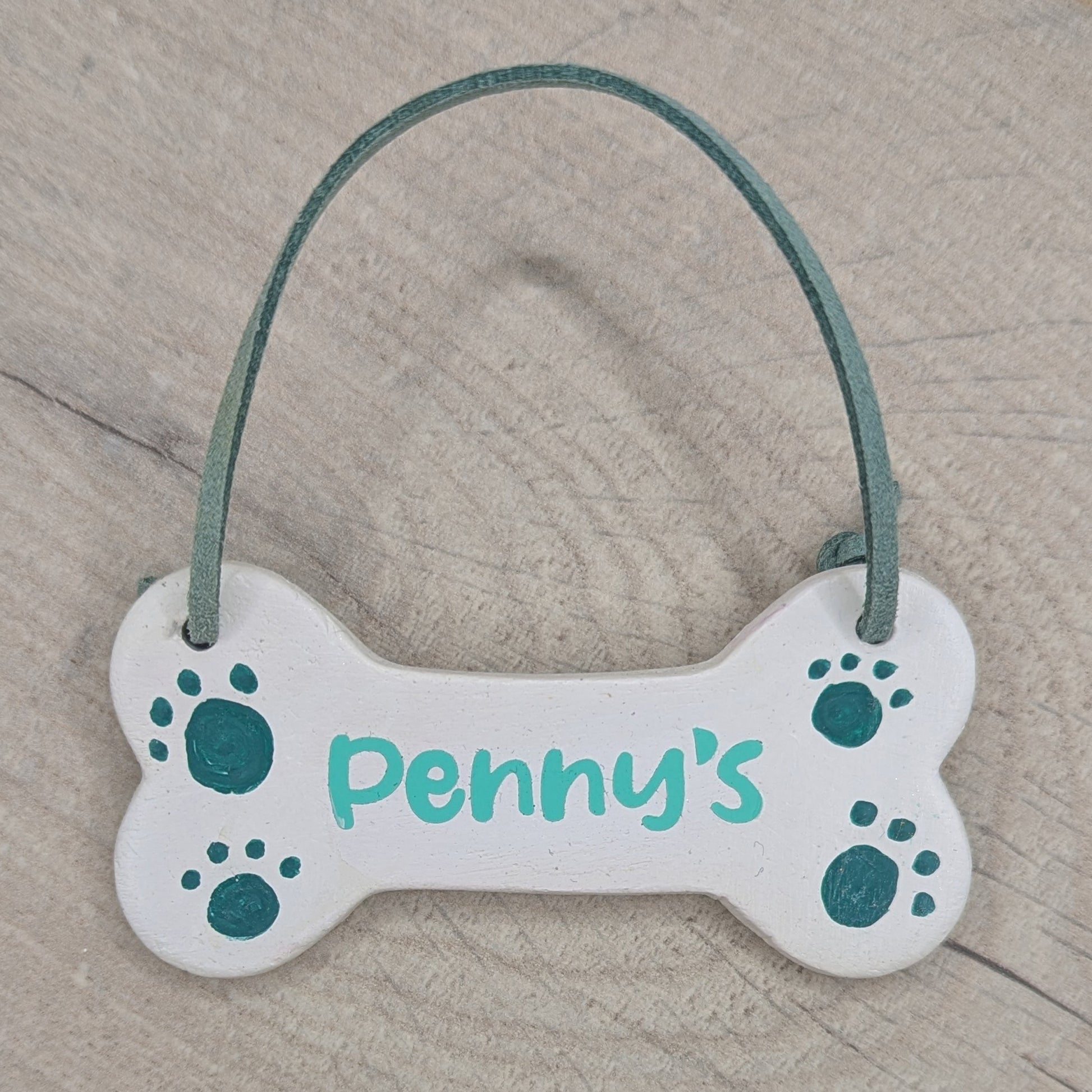decorated clay dog bone decoration, wording on it is the name Penny with 4 dog paw prints in green and a green leather hanging cord