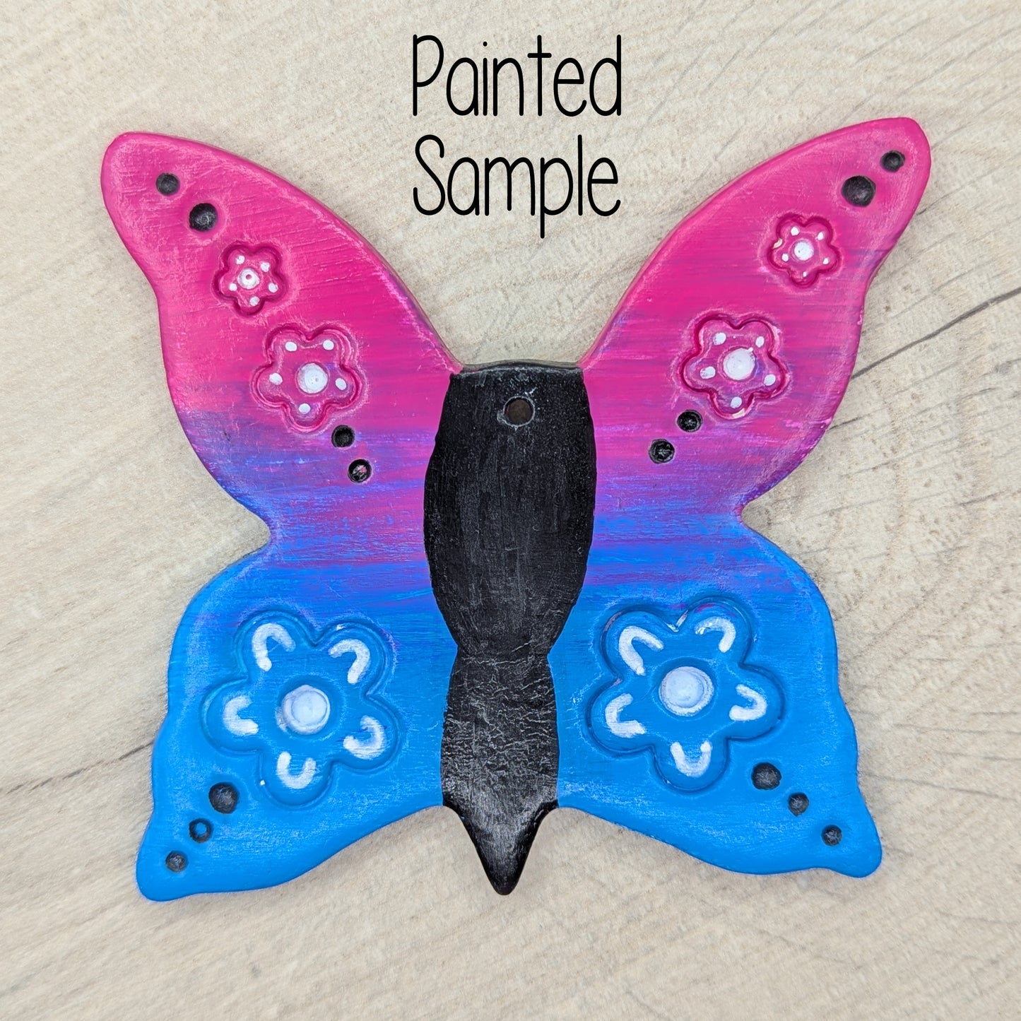 sample of large clay butterfly decoration, painted in bright pink and blue ombre effect, with a black body and white flowers on the wings