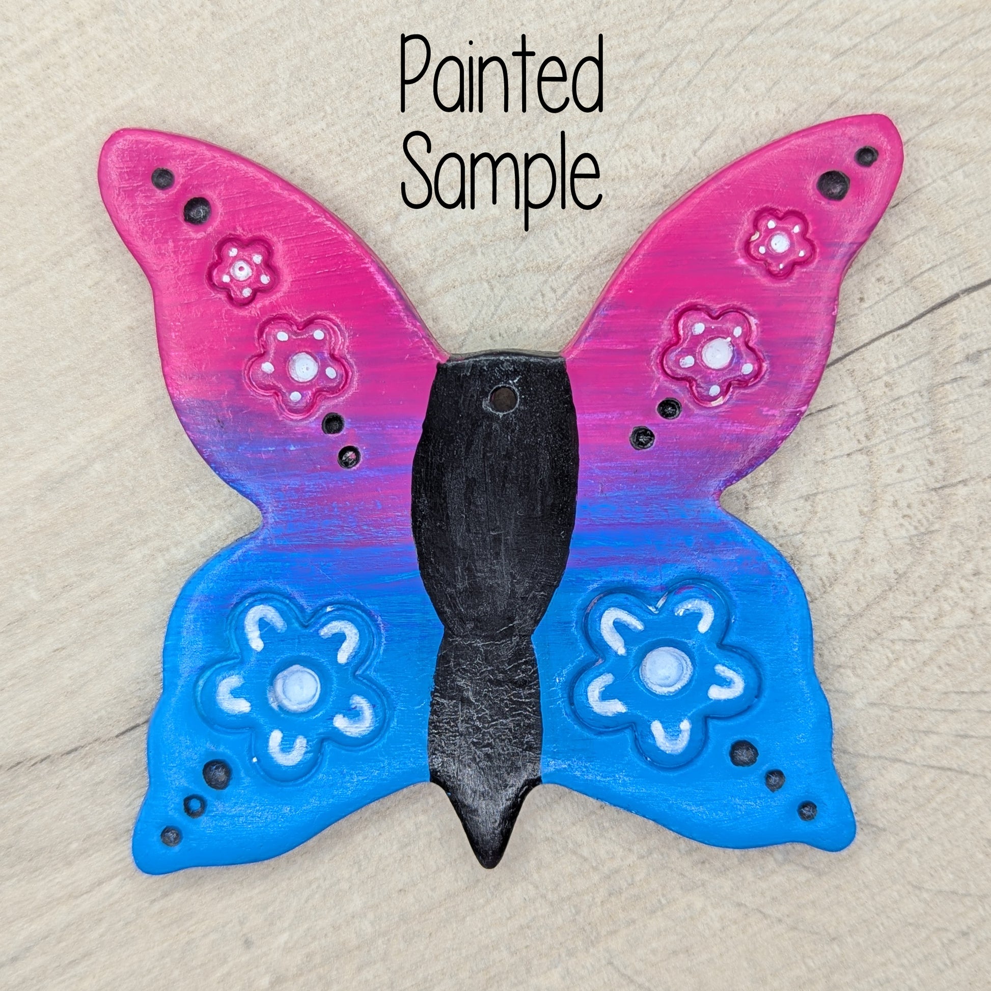 sample of large clay butterfly decoration, painted in bright pink and blue ombre effect, with a black body and white flowers on the wings
