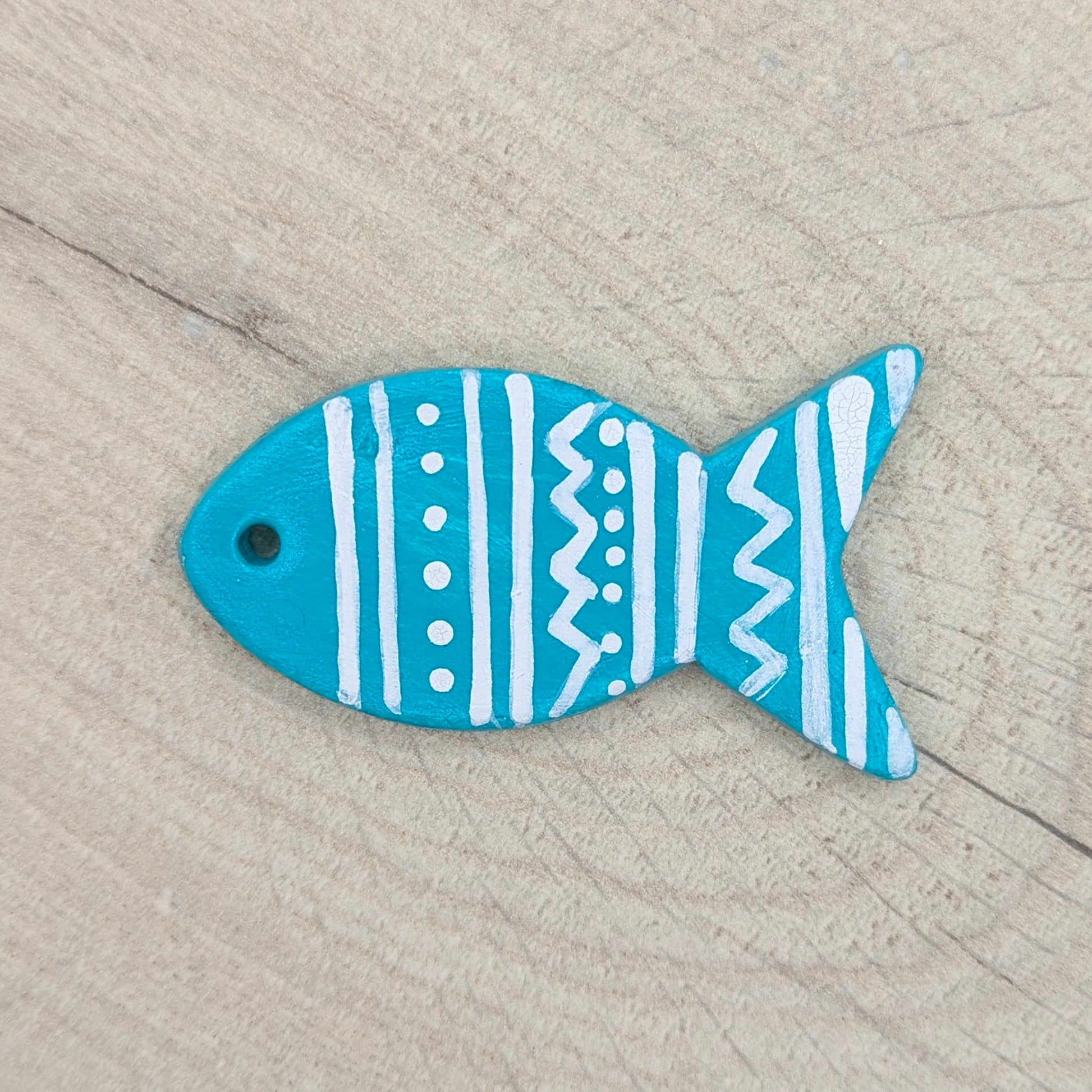small fish shaped clay decoration painted in mid blue with a white painted patten of spots and stripes, on a wooden background