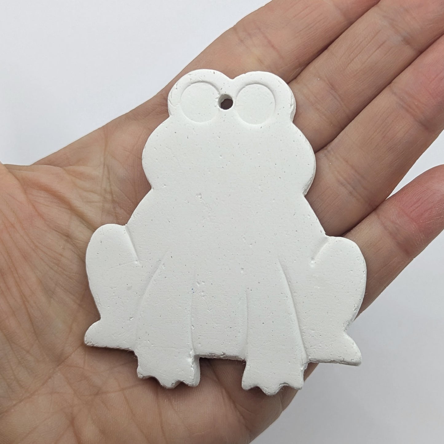 clay frog decoration made of white air dried clay, legs and eyes indented, shown in a ladies hand for scale