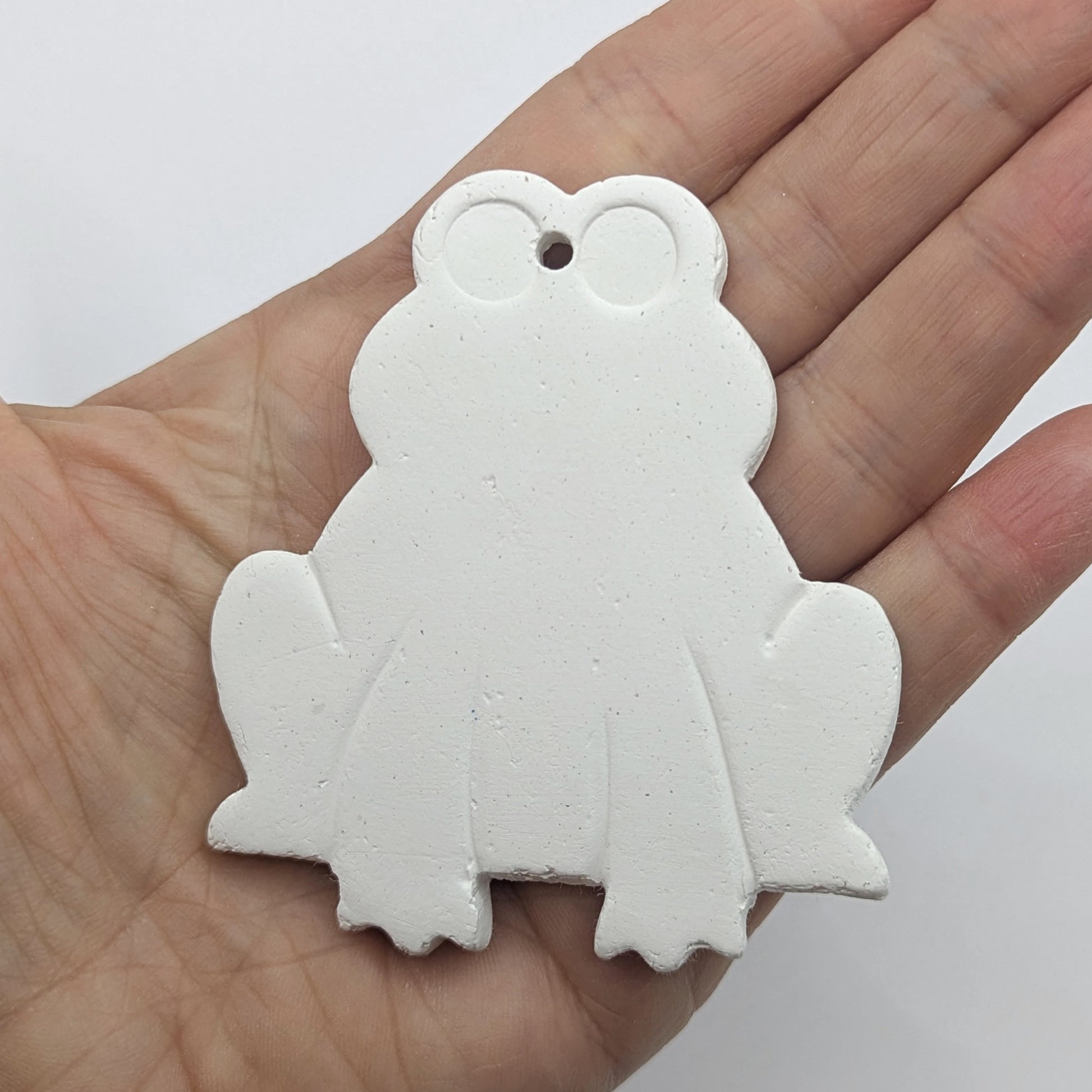 clay frog decoration made of white air dried clay, legs and eyes indented, shown in a ladies hand for scale