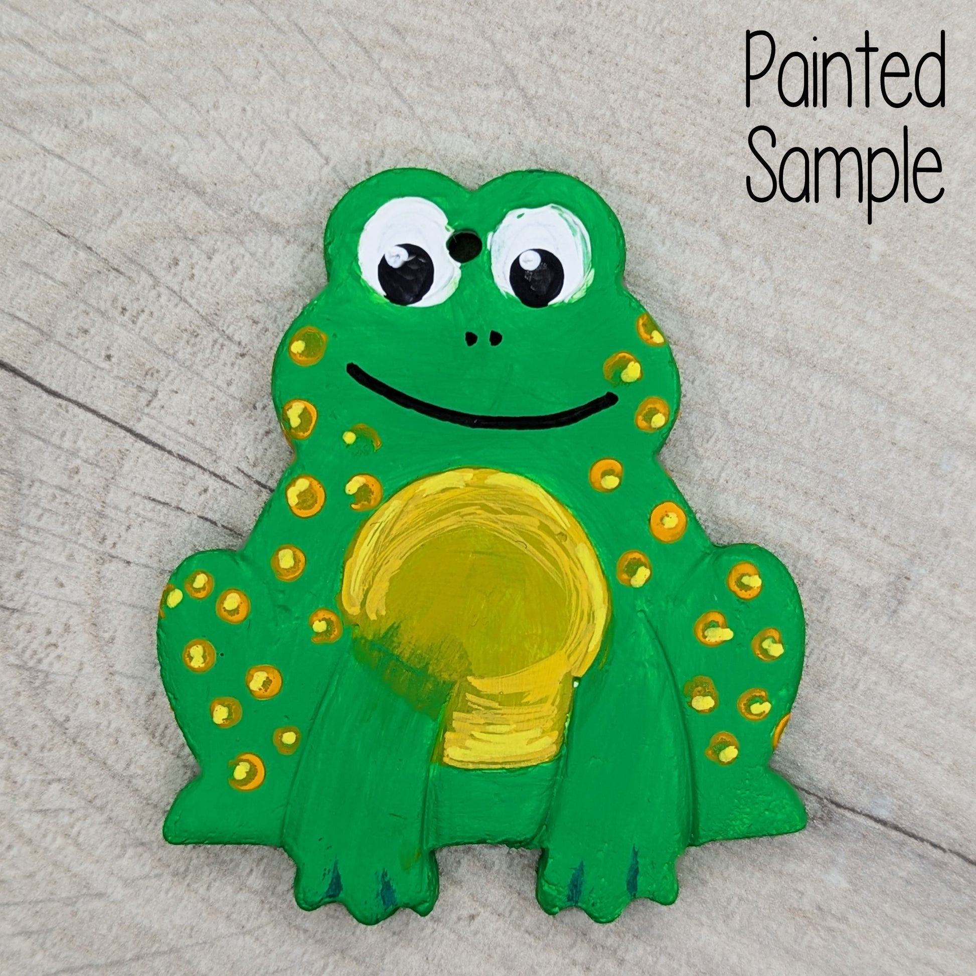 clay decoration painted in green and yellow acrylic to look like a frog, with a big smile and large eyes