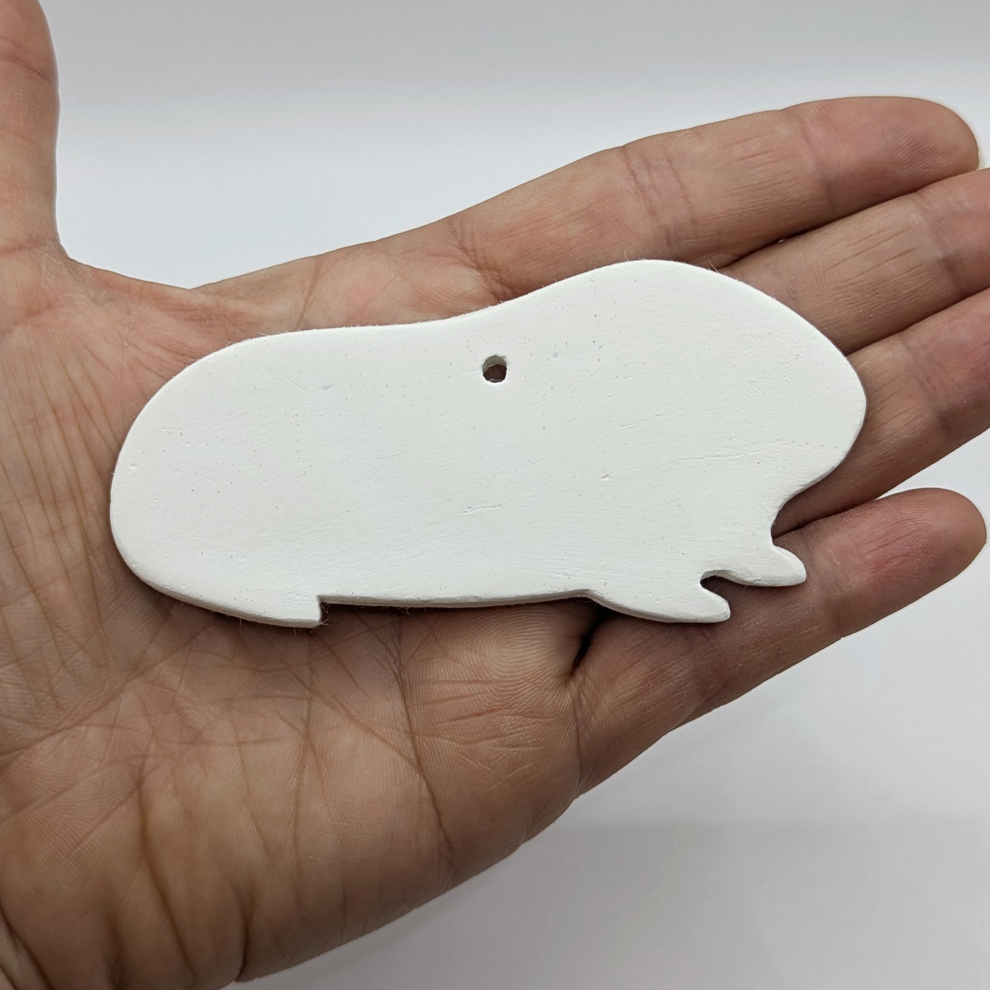 white air dried clay guinea pig shaped decoration on wooden background to paint at home, held in some ones hand to show size