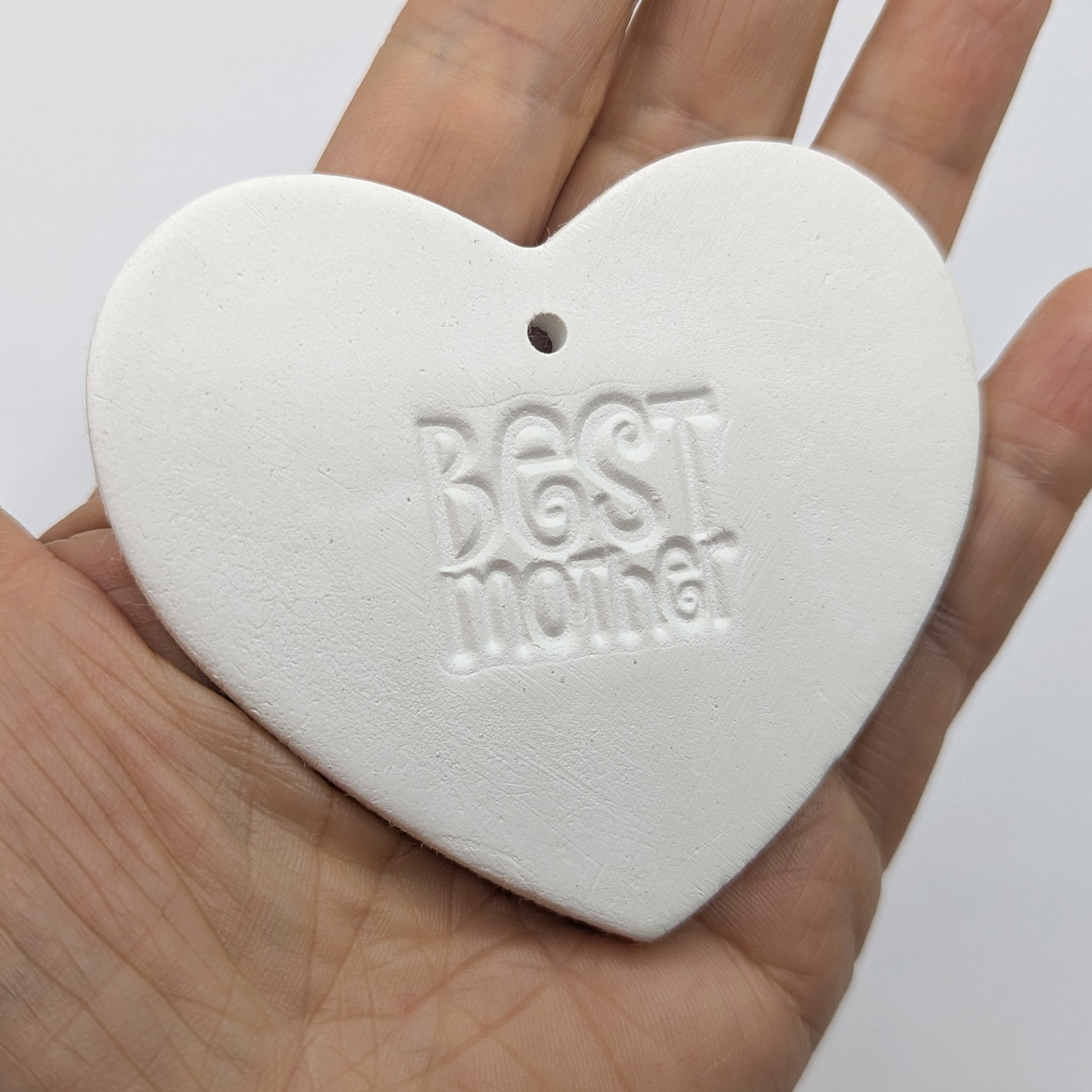 White heart shaped air dried clay tag with 'Best Mother' stamped into the clay, held in a hand against a white background