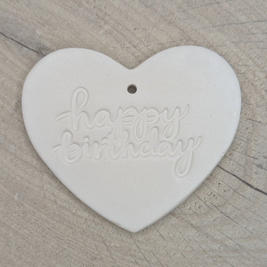 Heart shaped air dried clay decoration / tag with 'happy birthday' in lower case stamped into the clay, on a wooden background