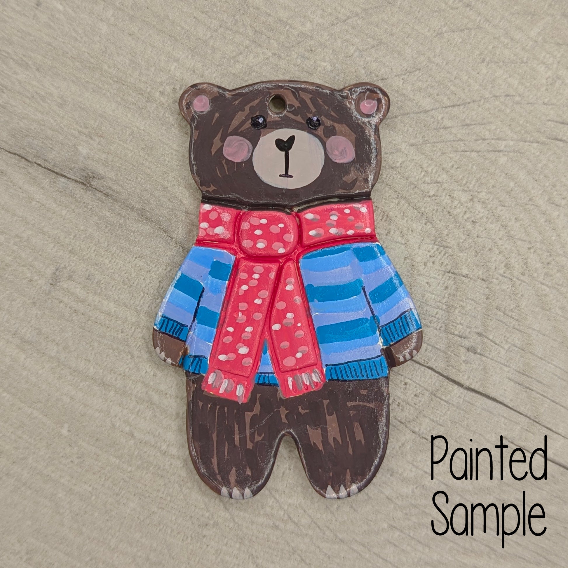 Painted bear ornament with a red scarf and blue striped sweater on a wooden surface, labeled 'Painted Sample'.
