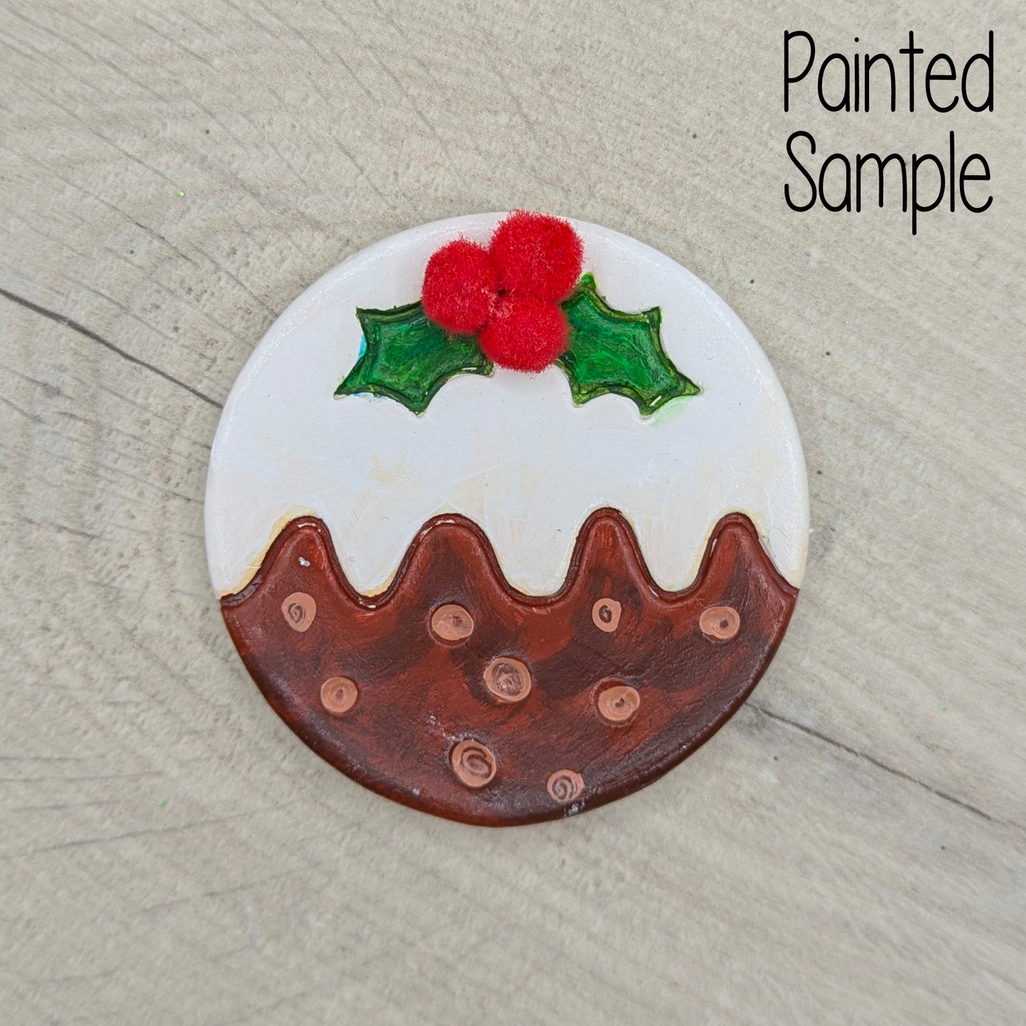 Painted decoration to look like a Christmas pudding with holly leaves and mini red pom poms for berries,  on a wooden surface