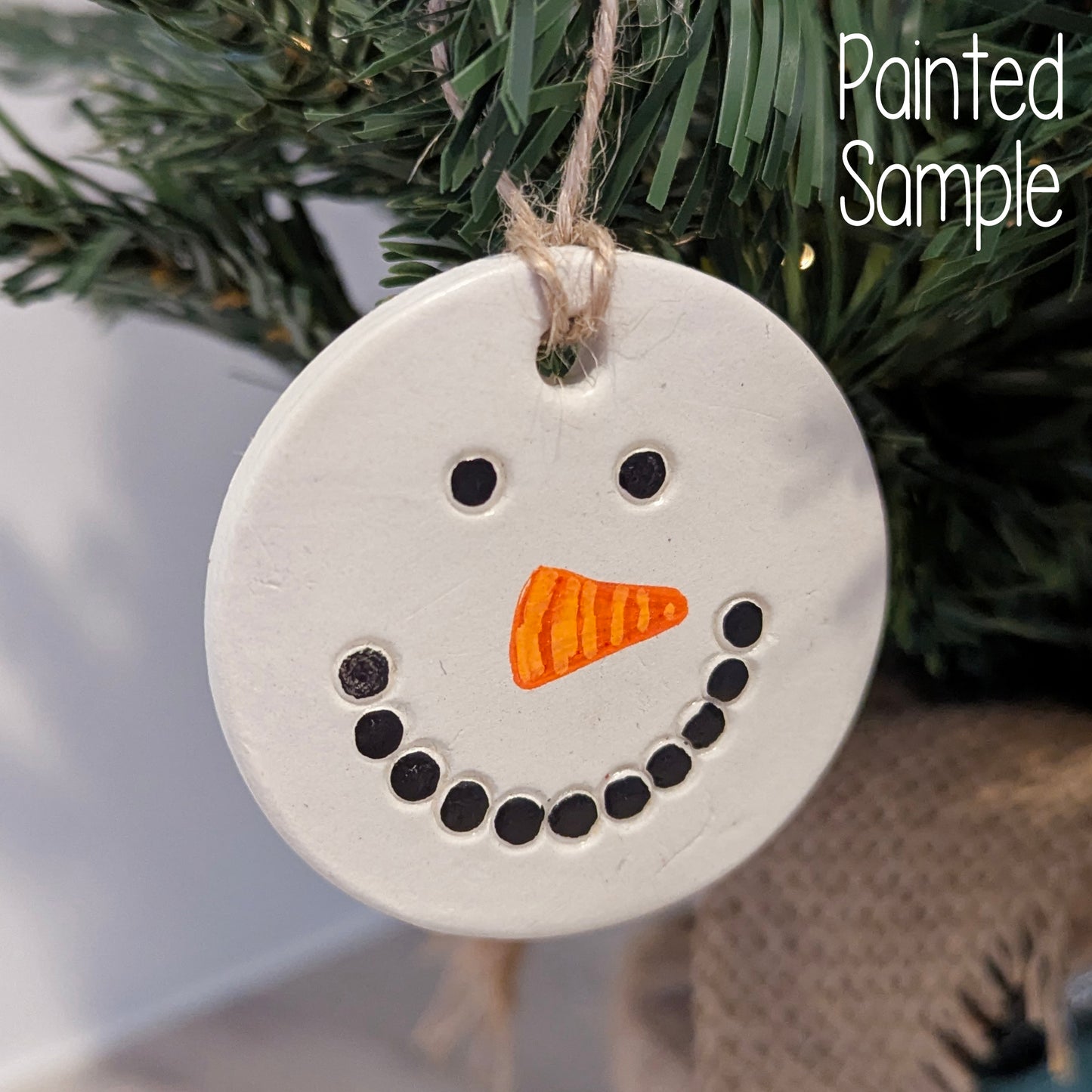 Round white ornament with black dots and orange carrot nose to represent a snowmans head, hanging on a tree.