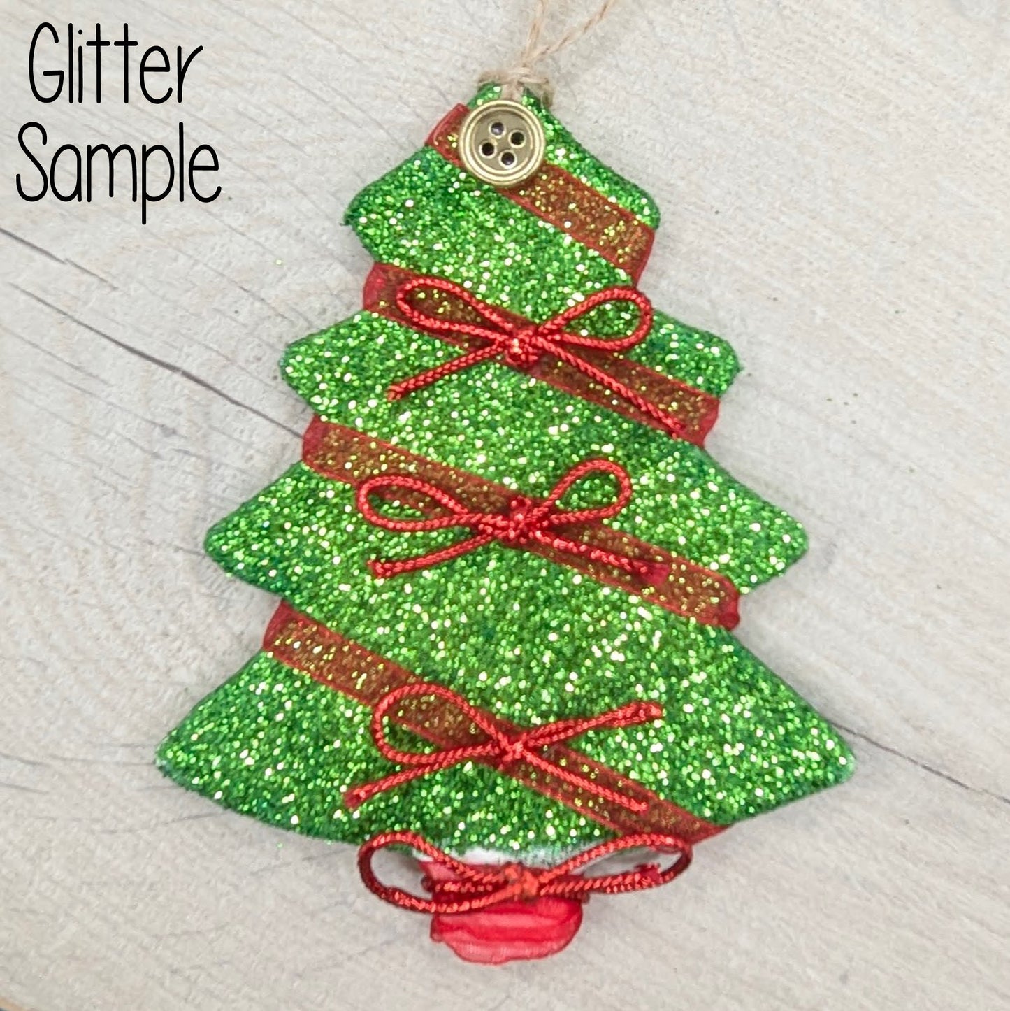 Green glitter Christmas tree decoration with red ribbons and a gold button on the top, displayed on a wooden surface.