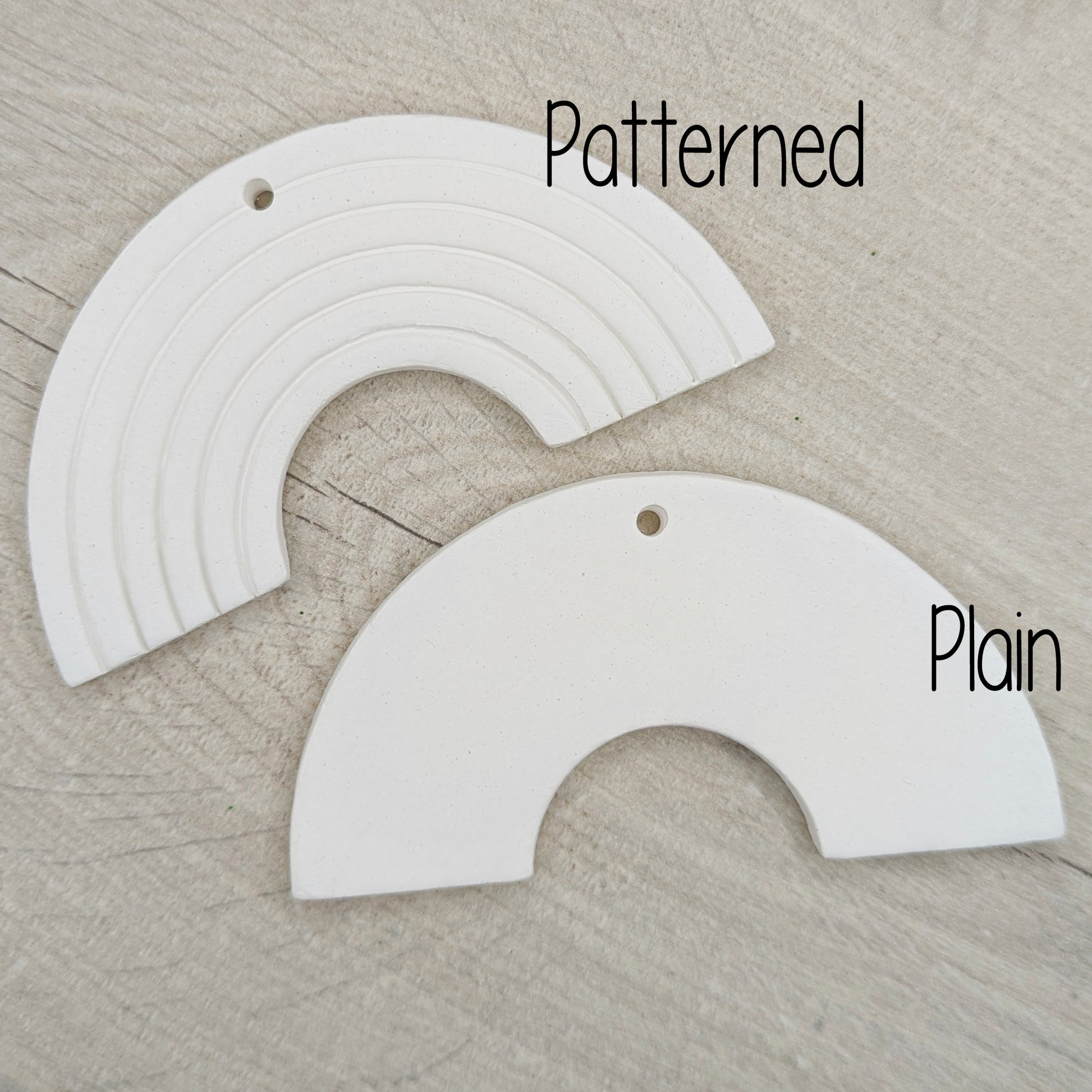 Two white clay rainbow arch shaped decorations, one patterned and one plain, on a wooden surface.