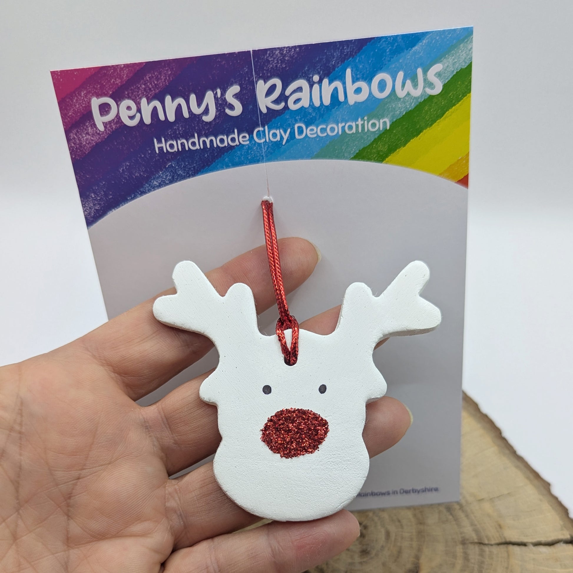 White clay reindeer ornament with red glitter nose and cord held in front of 'Penny's Rainbows' packaging