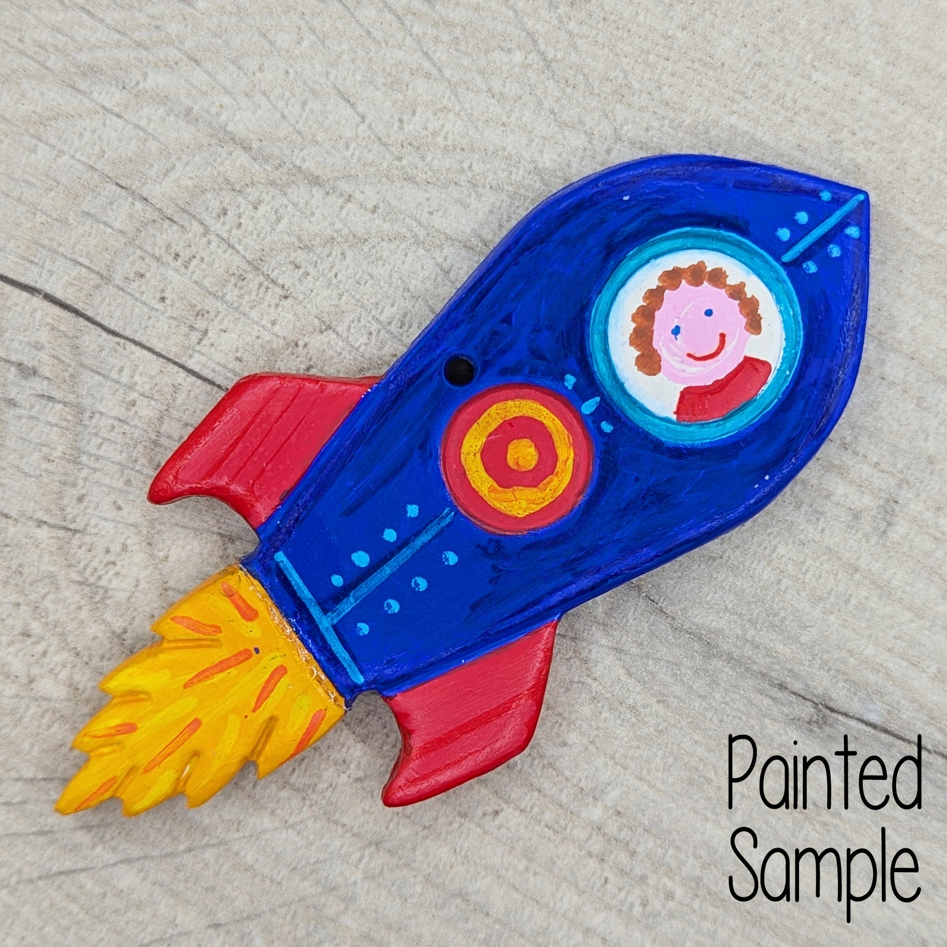painted rocket clay decoration, painted in blue, red and yellow with a smiley face looking through the window