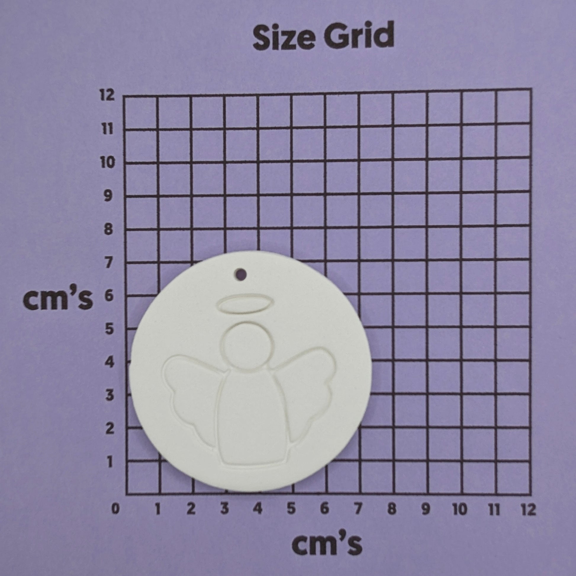 White clay disc with a simple clean angel design stamped onto it, shown on a purple cm size grid for scale