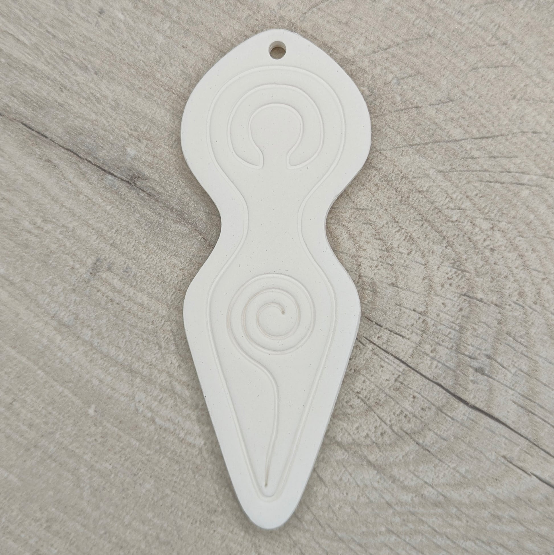 Spiral goddess shaped clay decoration with a hanging hole above her head area, shown on a light wood background