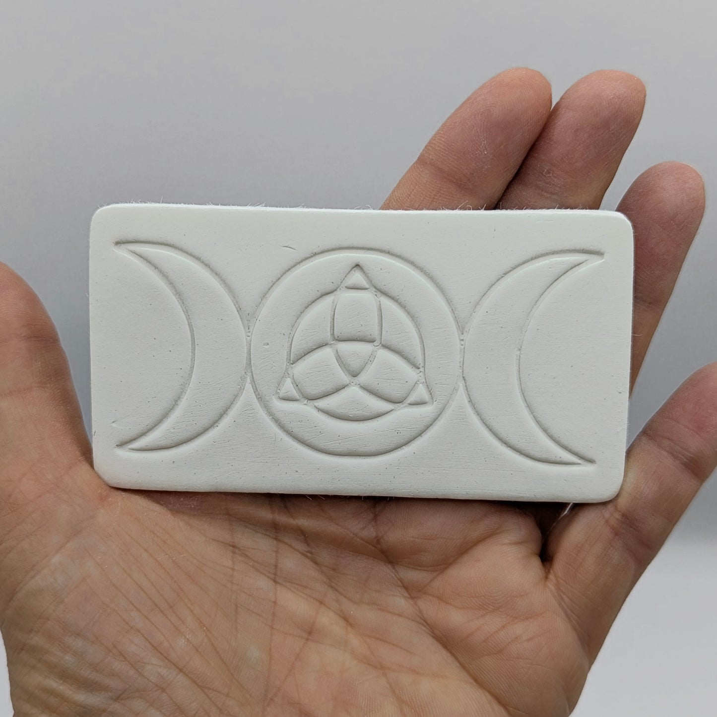 rectangular shaped air dried clay decoration with the triple moon and triquetra symbols stamped into it held in some ones hand
