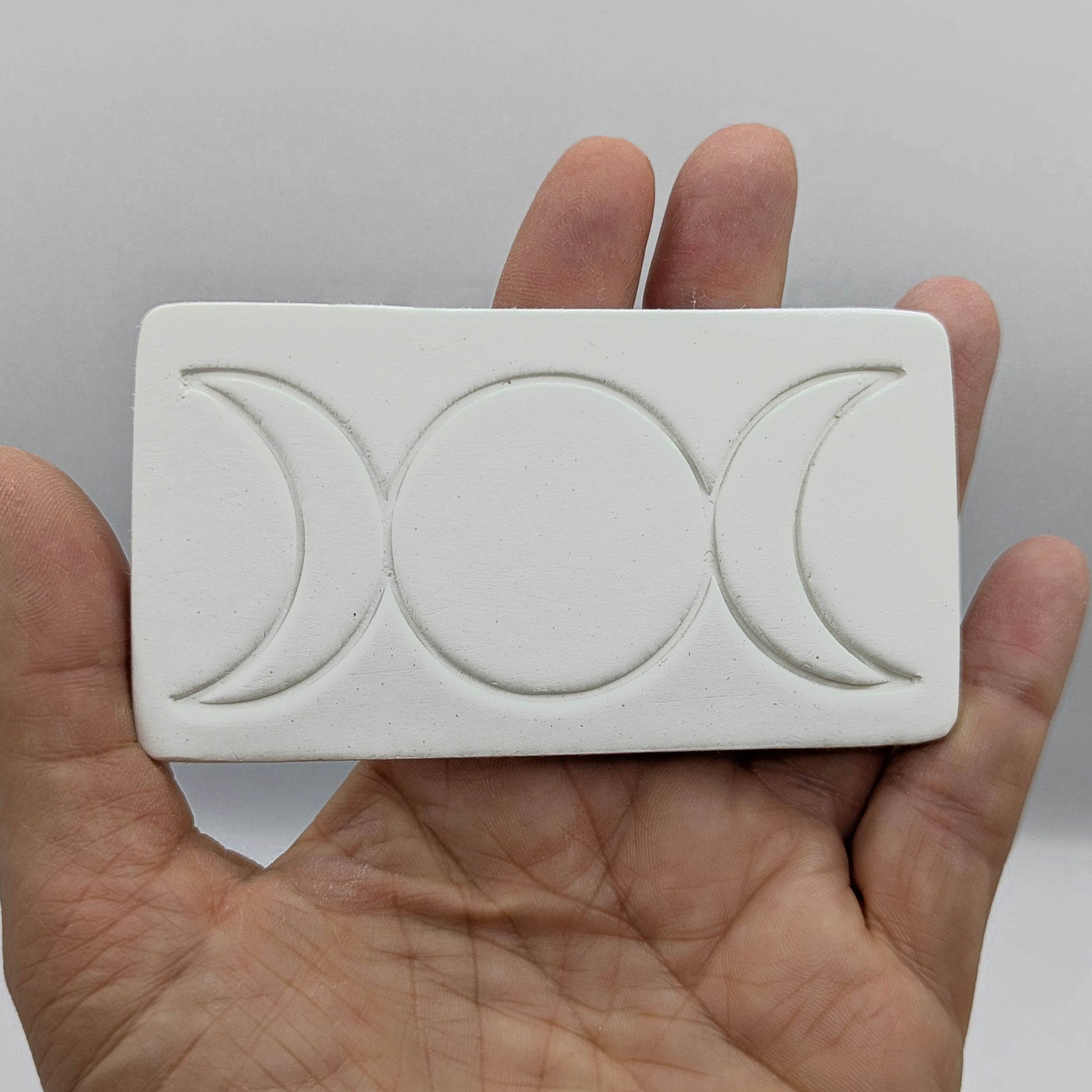 rectangular shaped air dried clay decoration with the triple moon symbol stamped into it held in some ones hand to show size