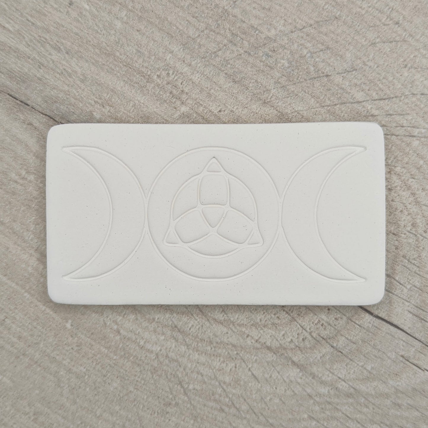 rectangular shaped air dried clay decoration with the triple moon and triquetra symbols stamped into it on a wooden background