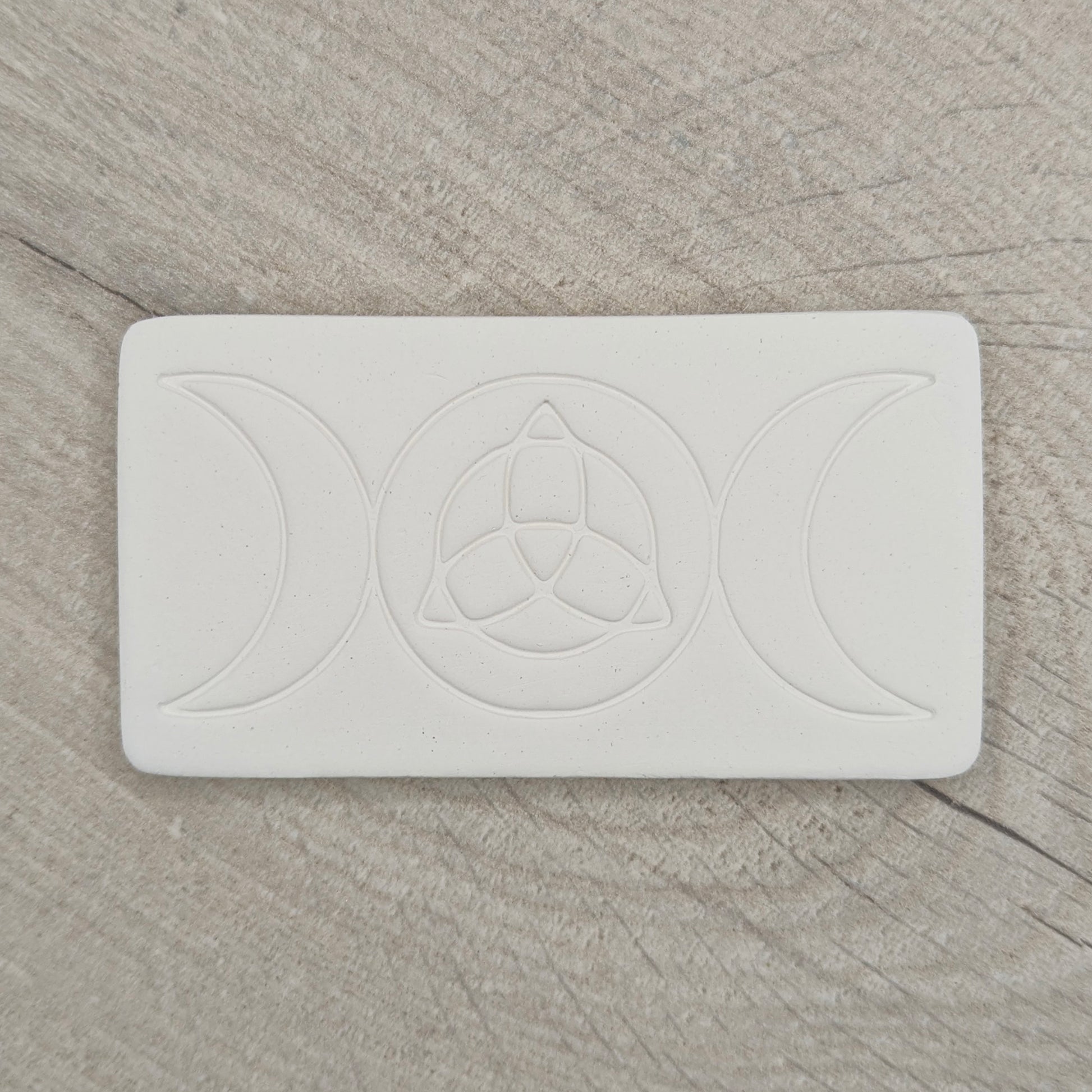 rectangular shaped air dried clay decoration with the triple moon and triquetra symbols stamped into it on a wooden background