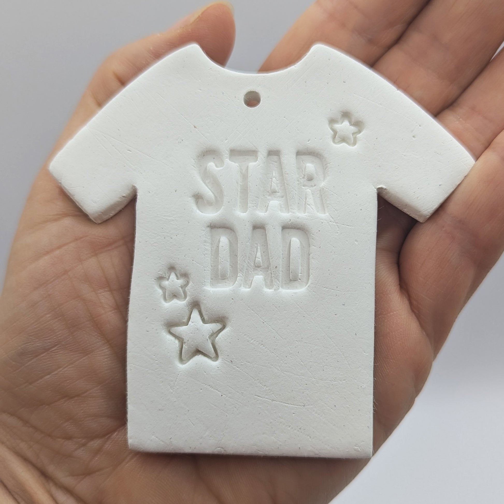 White clay t-shirt-shaped ornament / decoration with 'Star Dad' and small stars stamped into the clay, held in a hand to show scale
