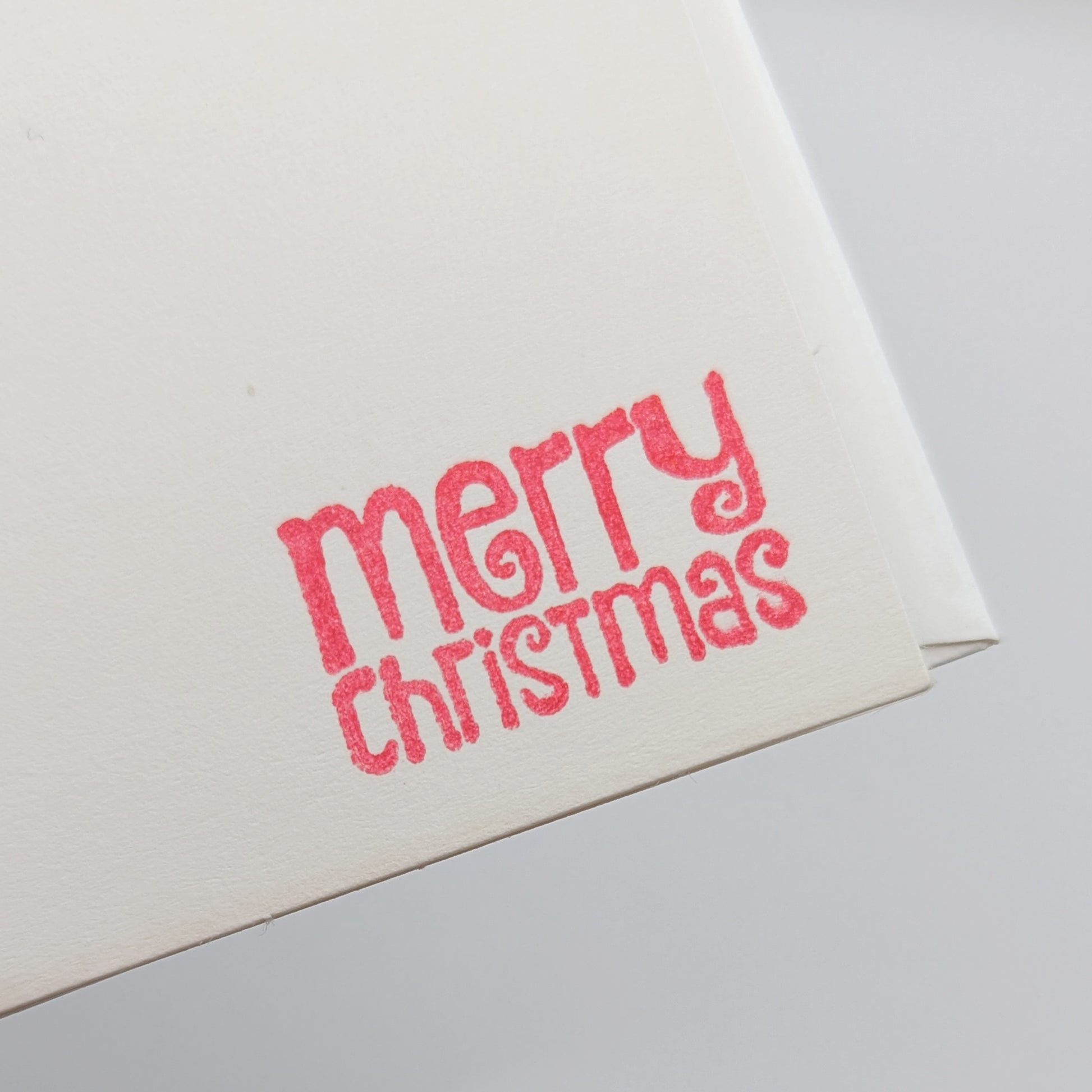close up of merry Christmas in a modern font, stamped in red ink onto a white card comes with matching envelope