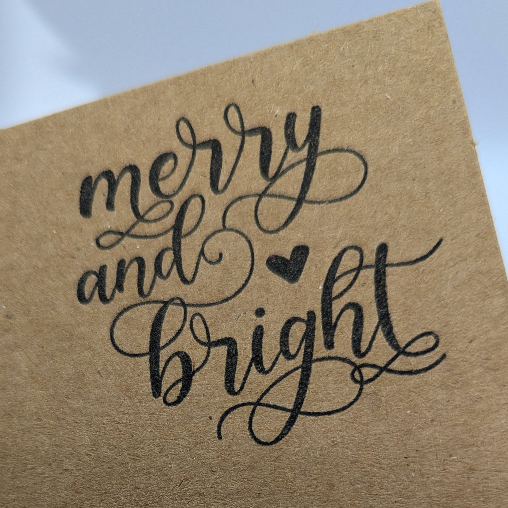 close up of wording 'merry and bright' in a script font, stamped in black ink onto a kraft card 