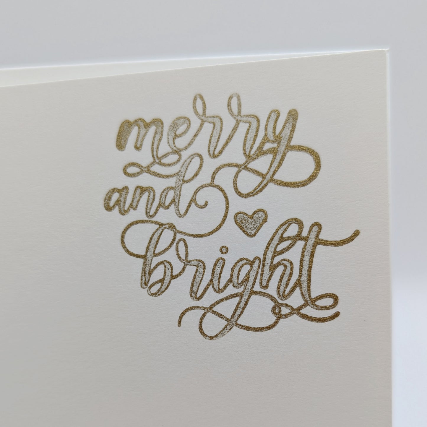 close up of wording 'merry and bright' in a script font, stamped in gold ink onto a white card 